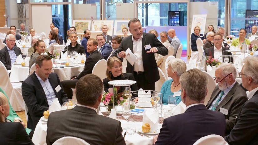 Mann spricht vor versammelter Gruppe bei formellem Dinner, Tische mit weißen Tischdecken, Gäste hören interessiert zu.