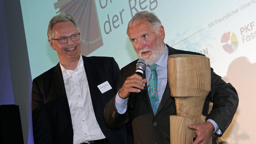 Zwei Männer bei einer Preisverleihung, einer hält eine Holzskulptur und ein Mikrofon, beide lächeln.