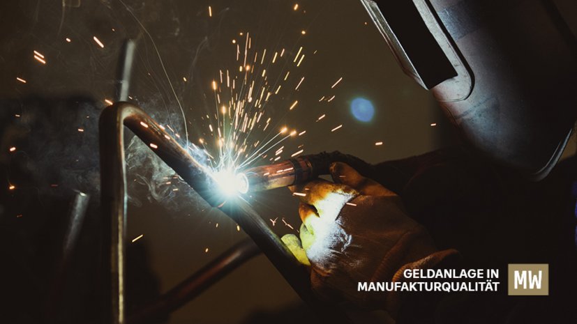 Arbeiter mit Schweißhelm schweißt Metall, Funken fliegen, Text: Geldanlage in Manufakturqualität, MW-Logo.