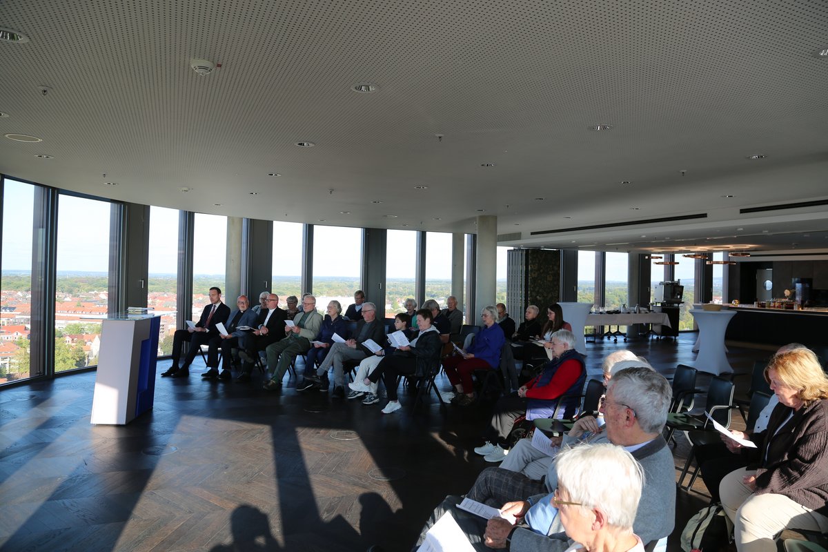 Gruppe von Personen in einem modernen Raum mit Panoramablick, die einer Präsentation aufmerksam folgen.