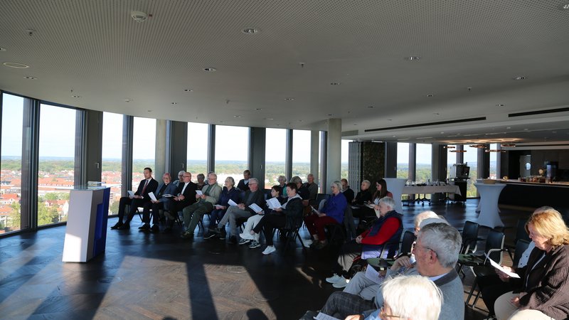 Gruppe von Personen in einem modernen Raum mit Panoramablick, die einer Präsentation aufmerksam folgen.