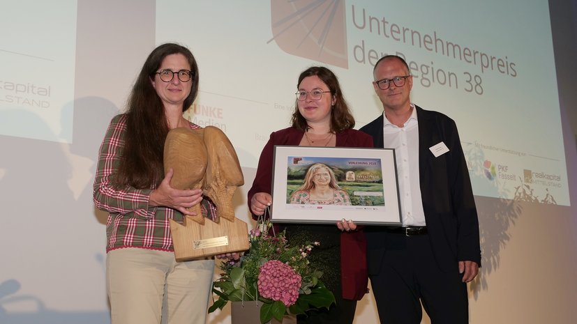 Drei Personen bei einer Preisverleihung; eine hält eine Trophäe, die andere eine gerahmte Urkunde und Blumen.