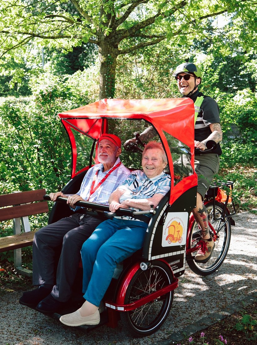Zwei Senioren genießen eine Rikscha-Fahrt durch einen sonnigen Park mit einem lachenden Fahrer.