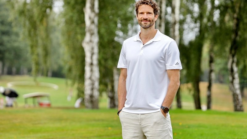 Mann (Arne Friedrich) steht in weißem Polohemd und hellen Hosen auf einem grünen Golfplatz, Bäume im Hintergrund.