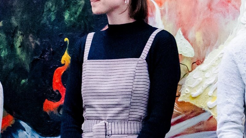 Frau steht vor farbenfrohem Wandgemälde, trägt schwarzen Pullover und kariertes Kleid, hält ein Buch in der Hand.