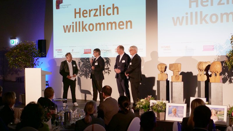 Vier Männer stehen auf einer Bühne mit Mikrofonen, vor einer Präsentation mit "Herzlich willkommen".