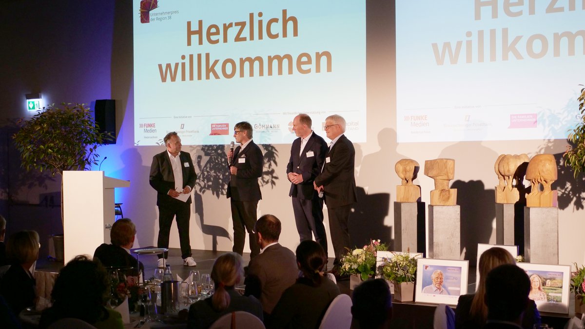 Vier Männer stehen auf einer Bühne mit Mikrofonen, vor einer Präsentation mit "Herzlich willkommen".
