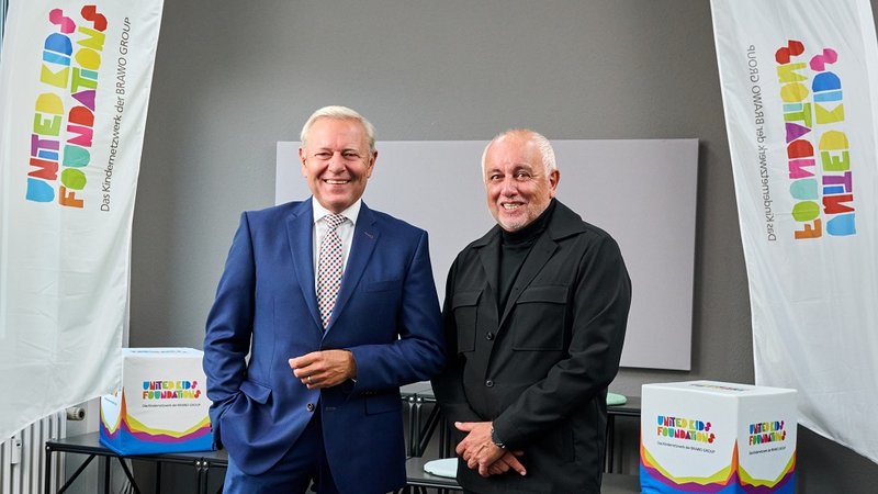 Zwei Männer in Anzügen stehen zwischen zwei Flaggen mit dem Logo der United Kids Foundation.
