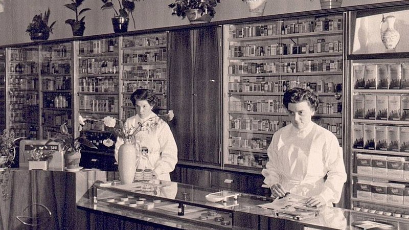 Zwei Frauen in einem historischen Apothekeninterieur mit Regalen voller Medikamente und Pflanzen.
