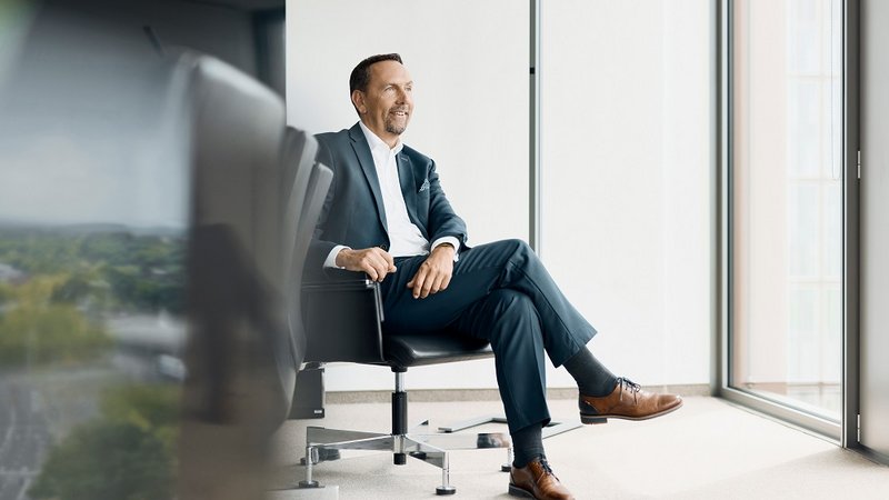 Ein Mann in einem eleganten Anzug sitzt entspannt auf einem Bürostuhl in einem modernen, hellen Raum. Der Raum hat große Fenster, die viel Tageslicht hineinlassen. Der Mann hat die Beine übereinandergeschlagen und lächelt in die Ferne. Seine braunen Lederschuhe und die dunklen Socken sind sichtbar. Links im Vordergrund ist ein unscharfer Konferenztisch zu sehen.