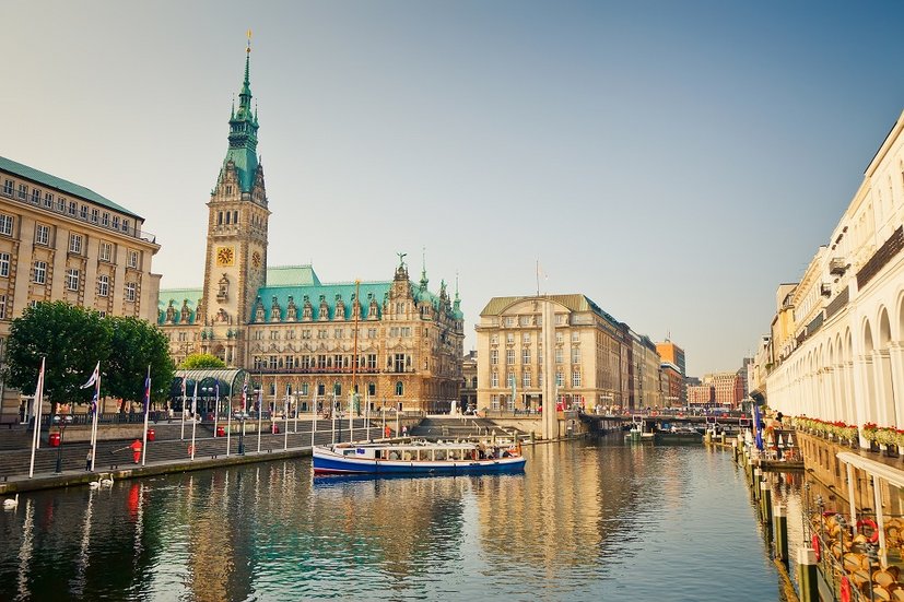Das Bild zeigt das historische Hamburger Rathaus am Rand eines ruhigen Kanals. Die Architektur des Rathauses ist beeindruckend mit einem hohen Turm und grünen Dächern. Im Vordergrund fährt ein Ausflugsboot auf dem Kanal, umgeben von eleganten Gebäuden und Bögen am Wasser. Flags und Bäume säumen die Uferpromenade unter einem klaren, blauen Himmel.