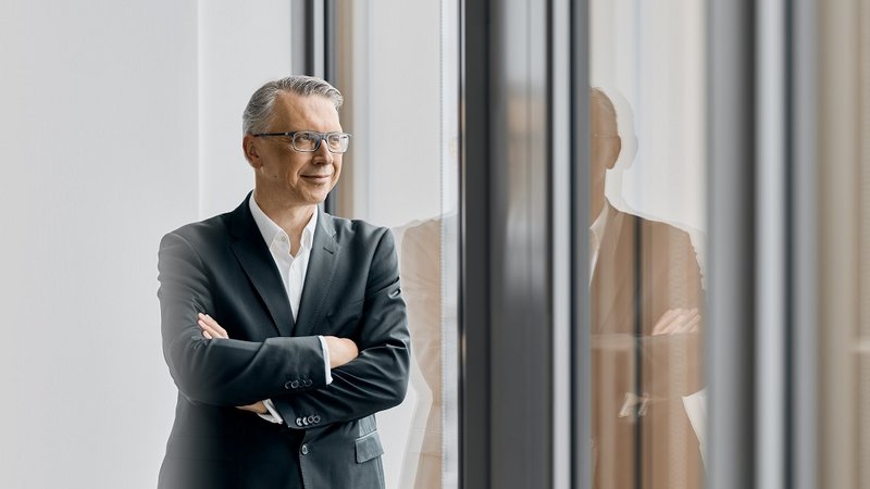 Mann im Anzug steht lächelnd vor einem Fenster, Arme verschränkt, mit natürlichem Licht und Fensterspiegelung.
