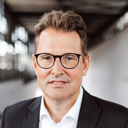 Mann in Anzug und Brille steht in einem modernen, lichtdurchfluteten Gebäude.