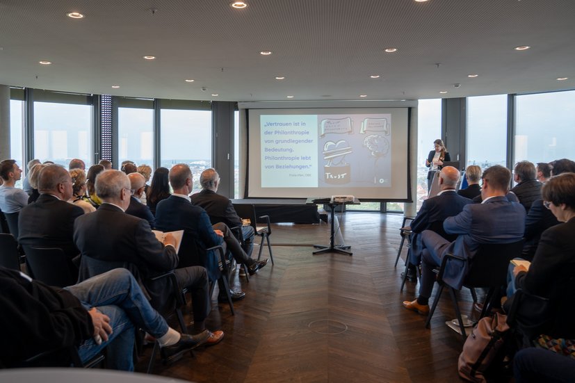 Person hält Präsentation in einem Konferenzraum, Zuschauer schauen auf eine Leinwand mit einer Folie über Vertrauen.