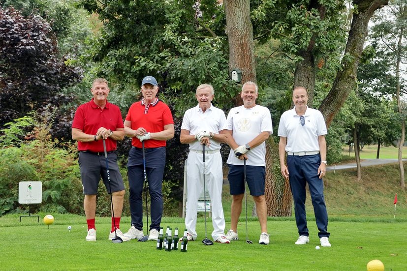 Fünf Männer stehen auf einem Golfplatz mit Golfschlägern, umgeben von Bäumen und Gras.
