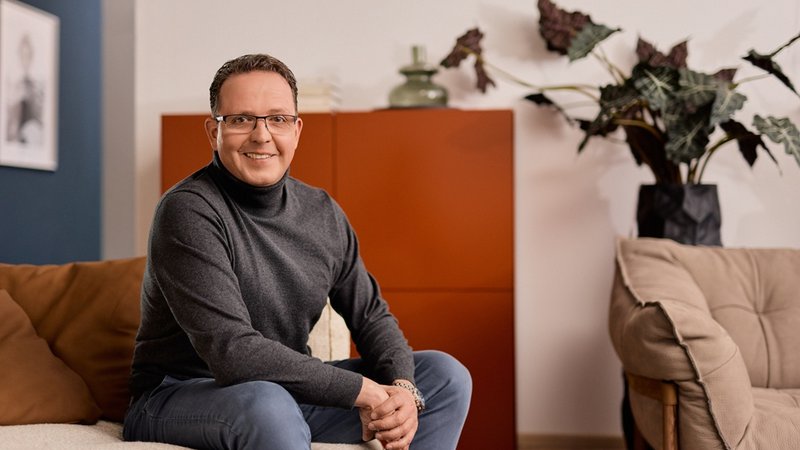 Mann mit Brille sitzt lächelnd auf Sofa, moderne Einrichtung und große Pflanze im Hintergrund.