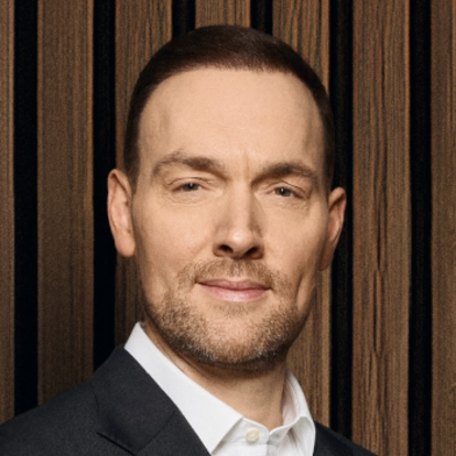 Mann in Anzug vor einem holzgetäfelten Hintergrund, mit neutralem Gesichtsausdruck.