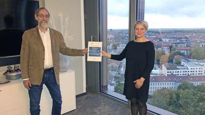 Zwei Personen überreichen ein Zertifikat in einem Büro mit Stadtblick im Hintergrund.