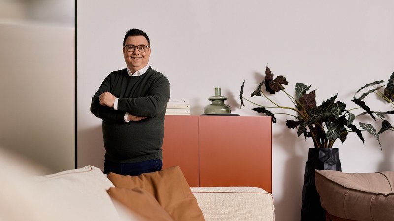 Mann mit Brille steht lächelnd in modernem Wohnzimmer mit Pflanzen und rotem Schrank im Hintergrund.