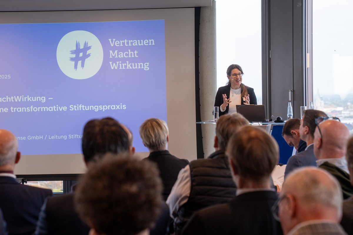 Frau hält einen Vortrag vor Publikum mit Projektorbildschirm "Vertrauen Macht Wirkung" im Hintergrund.