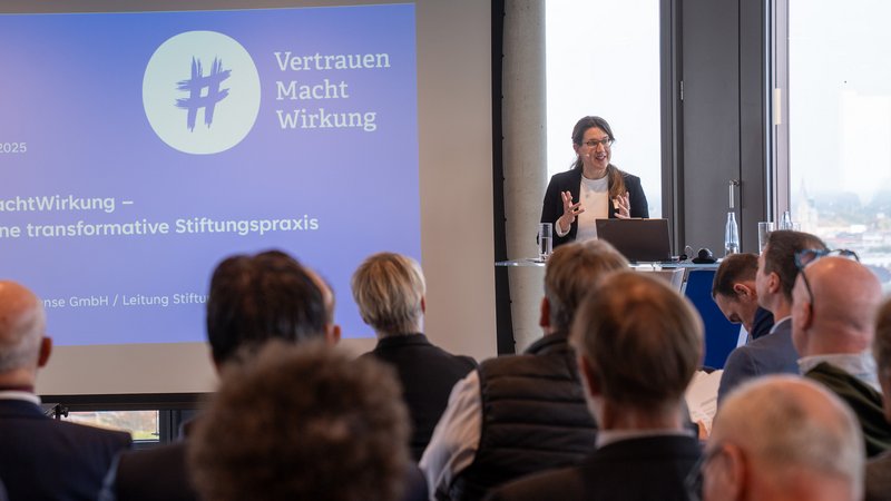 Frau hält einen Vortrag vor Publikum mit Projektorbildschirm "Vertrauen Macht Wirkung" im Hintergrund.