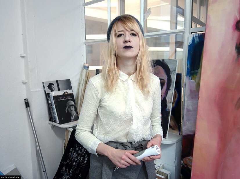 Person in weißer Bluse mit blonden Haaren steht in einem Atelier vor Kunstwerken und hält einen Gegenstand in der Hand.