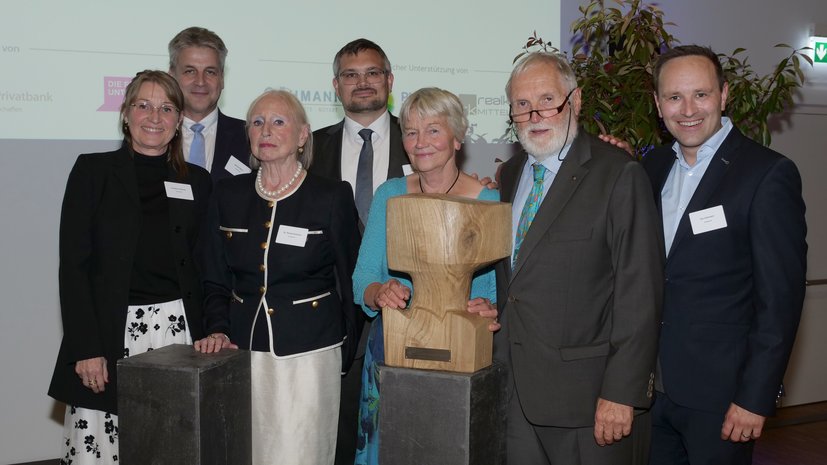 Eine Gruppe von sieben Personen posiert zusammen mit einer Holzskulptur bei einer Veranstaltung.