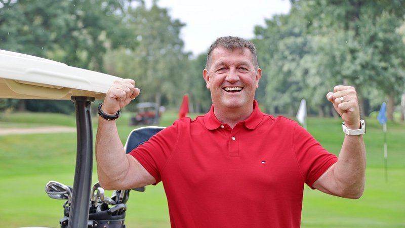 Mann in rotem Poloshirt lächelt und hebt die Arme freudig neben einem Golfwagen auf einem grünen Golfplatz.