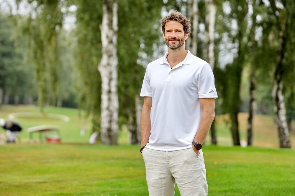 Mann (Arne Friedrich) steht in weißem Polohemd und hellen Hosen auf einem grünen Golfplatz, Bäume im Hintergrund.