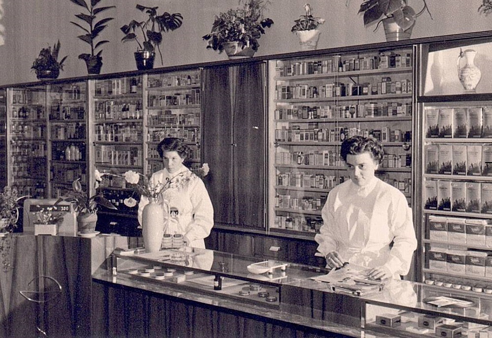Zwei Frauen in einem historischen Apothekeninterieur mit Regalen voller Medikamente und Pflanzen.