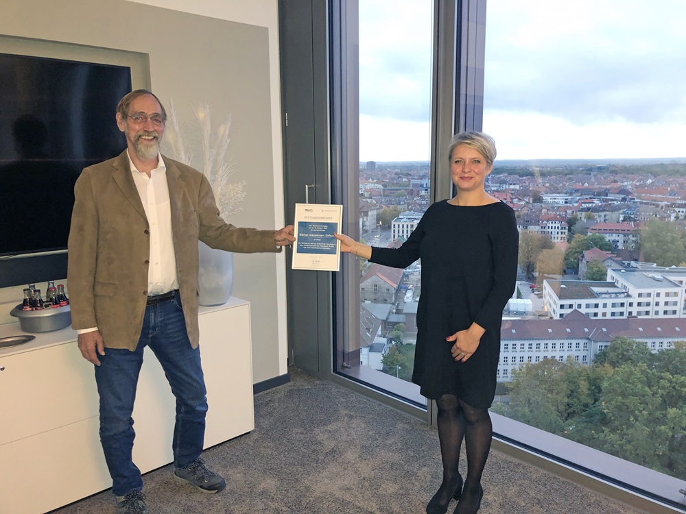Zwei Personen überreichen ein Zertifikat in einem Büro mit Stadtblick im Hintergrund.