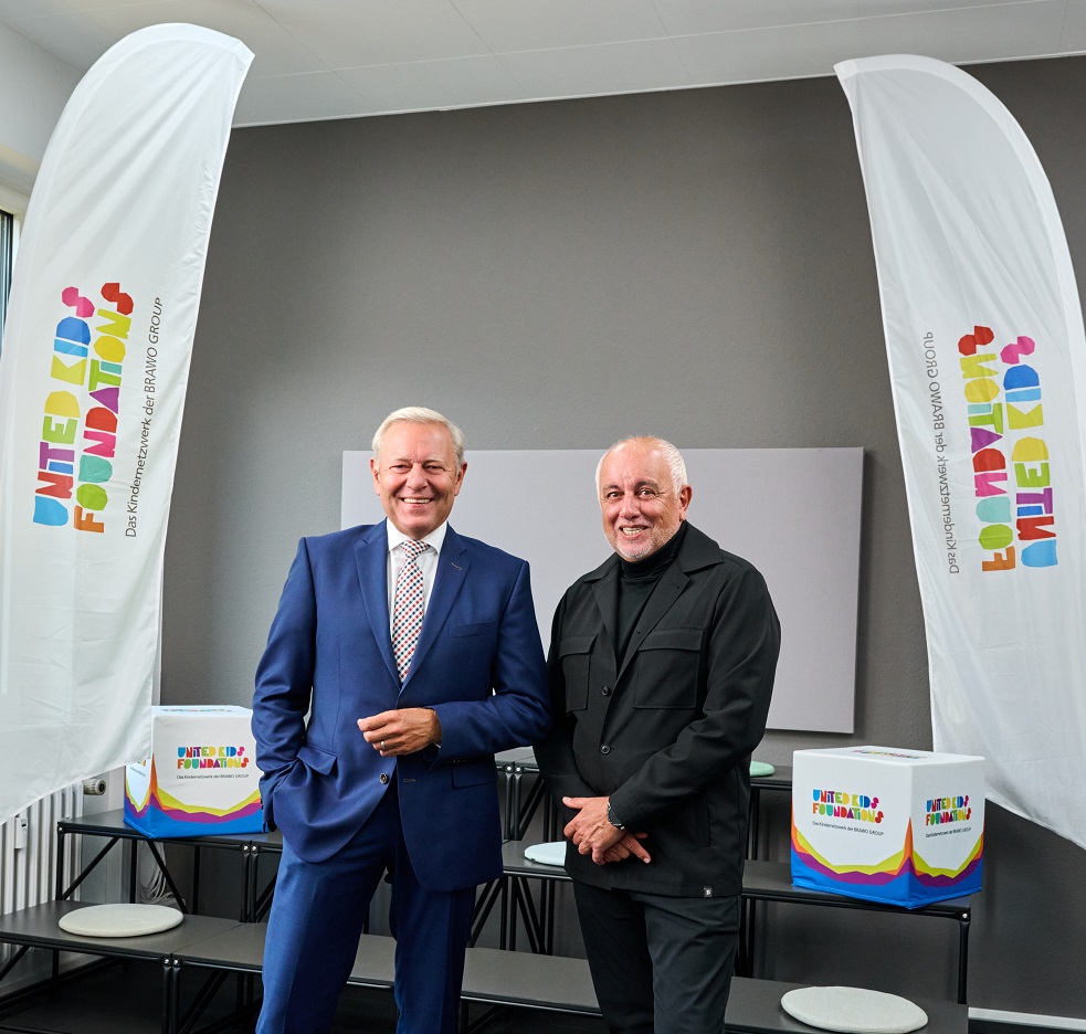 Zwei Männer in Anzügen stehen zwischen zwei Flaggen mit dem Logo der United Kids Foundation.