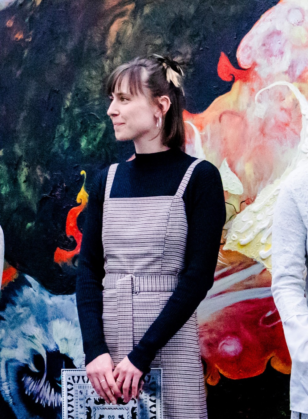 Frau steht vor farbenfrohem Wandgemälde, trägt schwarzen Pullover und kariertes Kleid, hält ein Buch in der Hand.