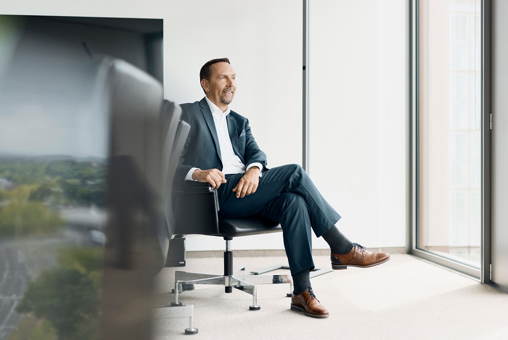 Ein Mann in einem eleganten Anzug sitzt entspannt auf einem Bürostuhl in einem modernen, hellen Raum. Der Raum hat große Fenster, die viel Tageslicht hineinlassen. Der Mann hat die Beine übereinandergeschlagen und lächelt in die Ferne. Seine braunen Lederschuhe und die dunklen Socken sind sichtbar. Links im Vordergrund ist ein unscharfer Konferenztisch zu sehen.