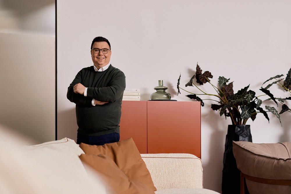 Mann mit Brille steht lächelnd in modernem Wohnzimmer mit Pflanzen und rotem Schrank im Hintergrund.