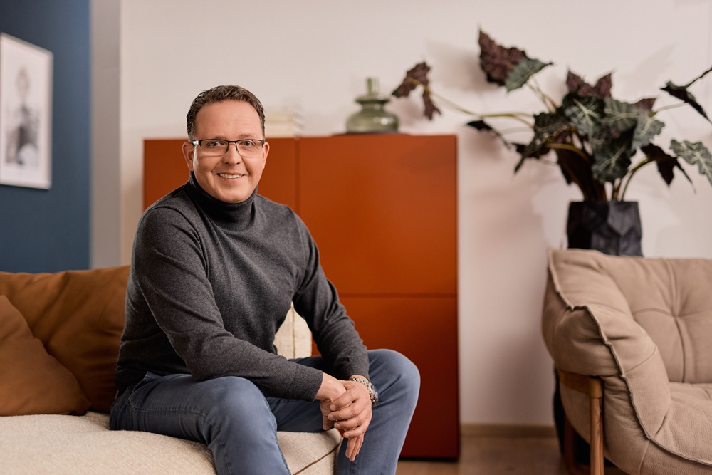 Mann mit Brille sitzt lächelnd auf Sofa, moderne Einrichtung und große Pflanze im Hintergrund.