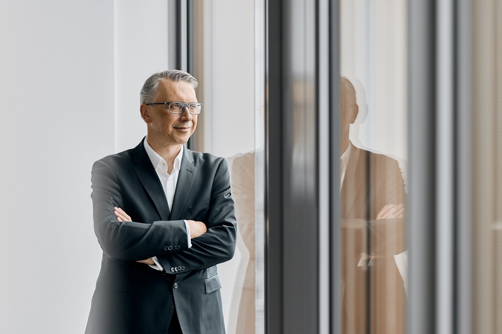 Mann im Anzug steht lächelnd vor einem Fenster, Arme verschränkt, mit natürlichem Licht und Fensterspiegelung.